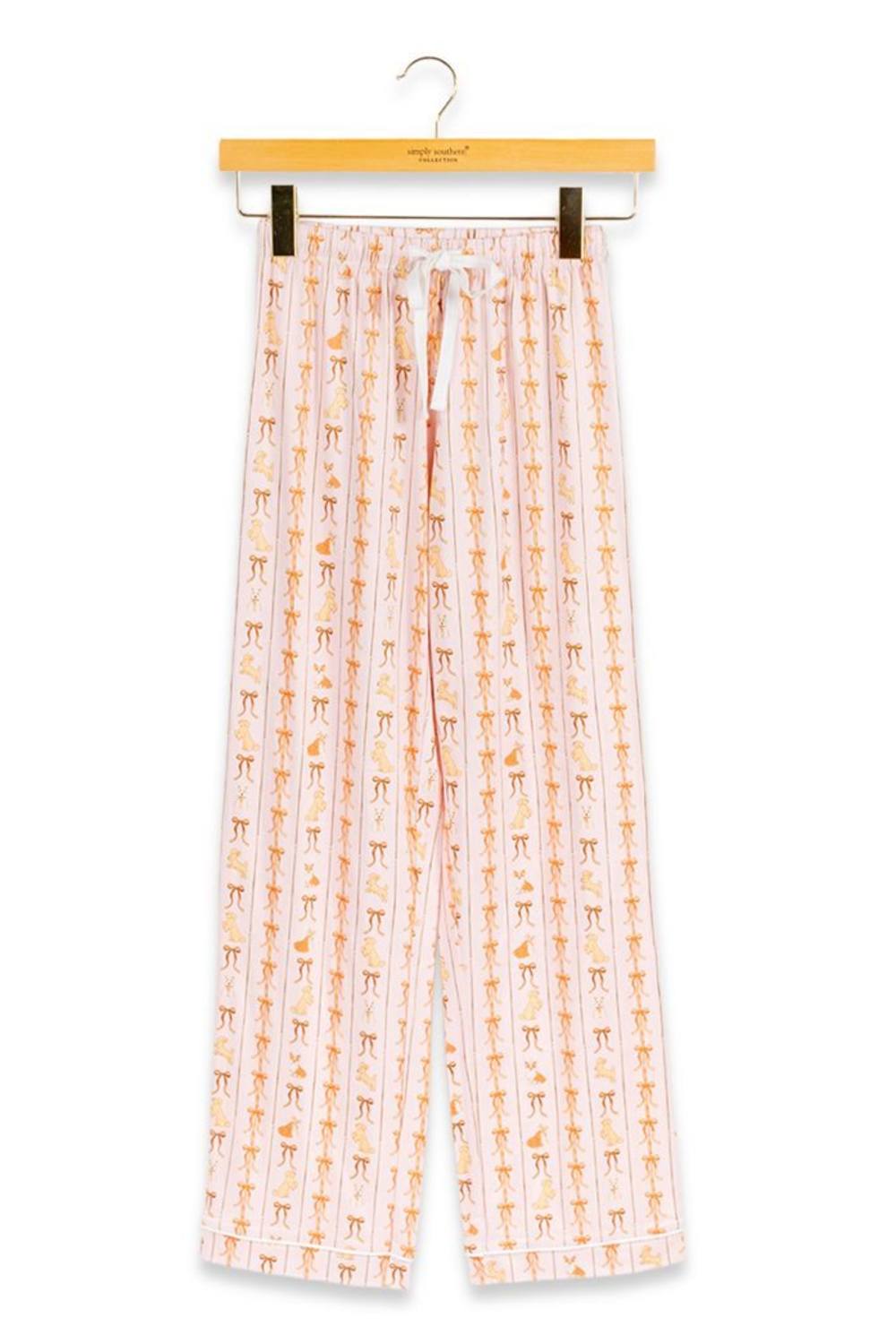 A pair of children's pajama pants hanging from a wooden hanger against a plain white background. The pants are light pink with a vertical pattern of small orange and yellow cartoon figures, and have a white drawstring waist.