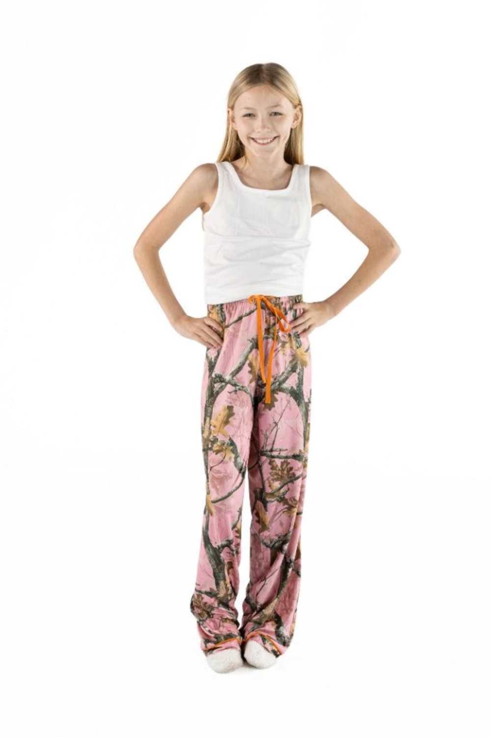 A young girl stands against a white background wearing a white tank top and pink camouflage print pajama pants with an orange drawstring.