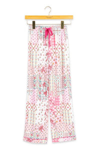 A pair of wide-leg pajama pants with a pink and white patchwork bohemian pattern, hanging on a wooden hanger against a white background.