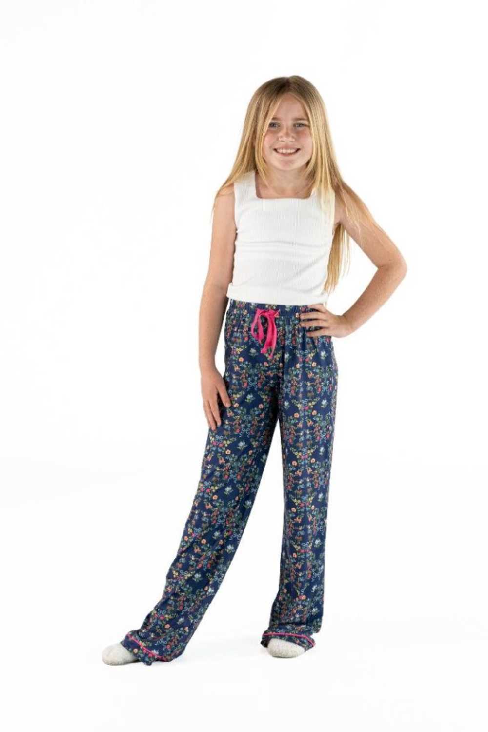 A young girl with long blonde hair stands against a white studio background wearing a white sleeveless top and dark blue floral pajama pants with a pink drawstring.