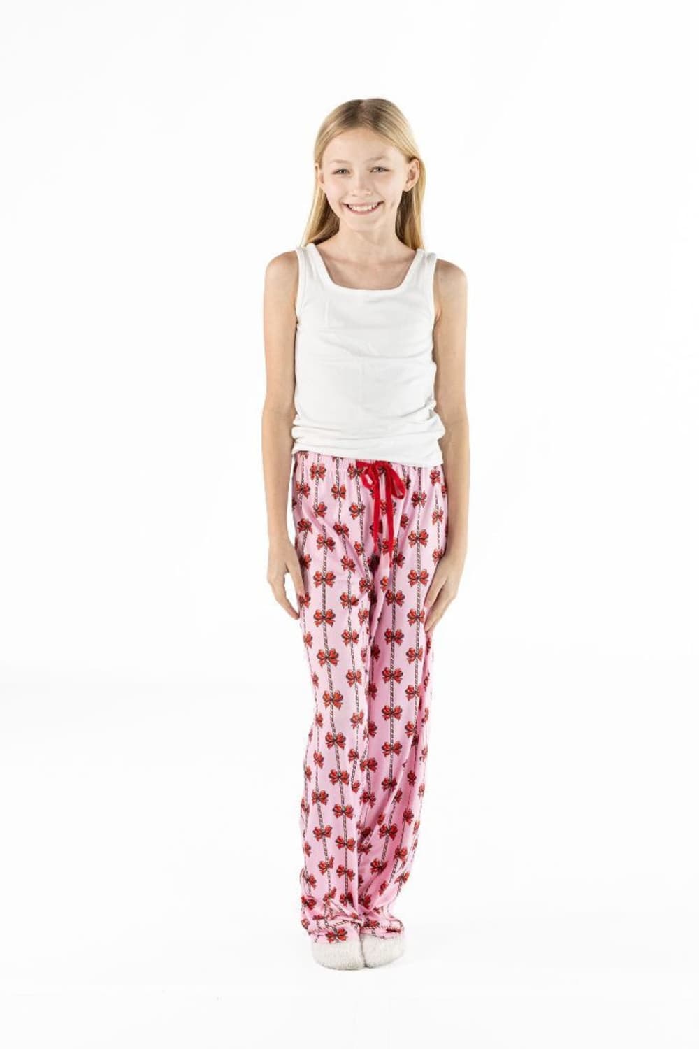 A young girl stands against a white studio background wearing a white sleeveless top and pink pajama pants with a red heart pattern and a red drawstring.