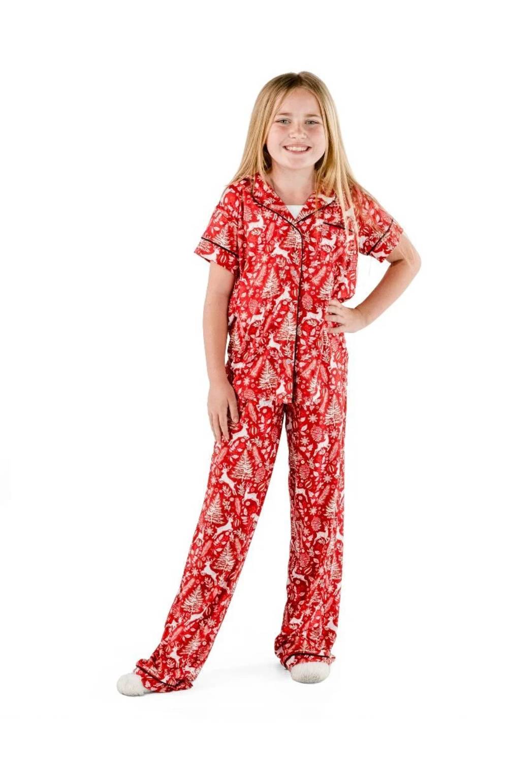 A young girl with blonde hair stands against a white background wearing a red short-sleeve pajama set with a white Christmas tree pattern.