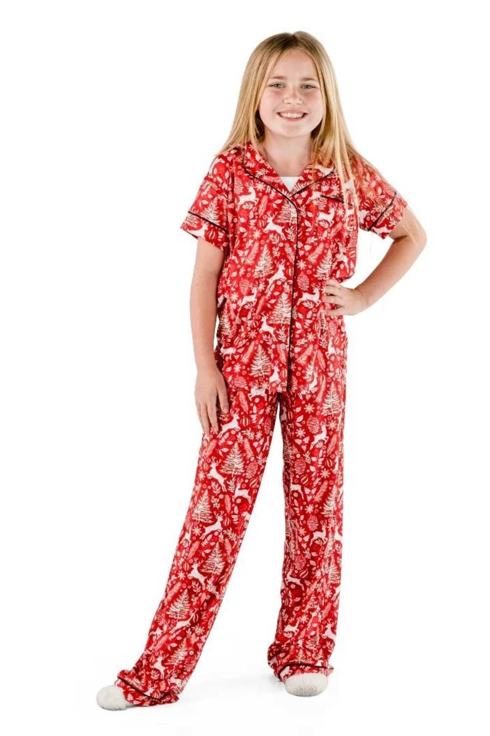 A young girl modeling a red short-sleeve pajama set with a white Christmas tree and deer pattern, standing against a plain white background.