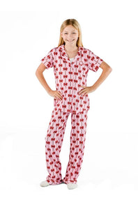 A young girl with blonde hair stands wearing a matching short-sleeve pajama set with a red bow pattern on a white background, posed against a solid white studio backdrop.