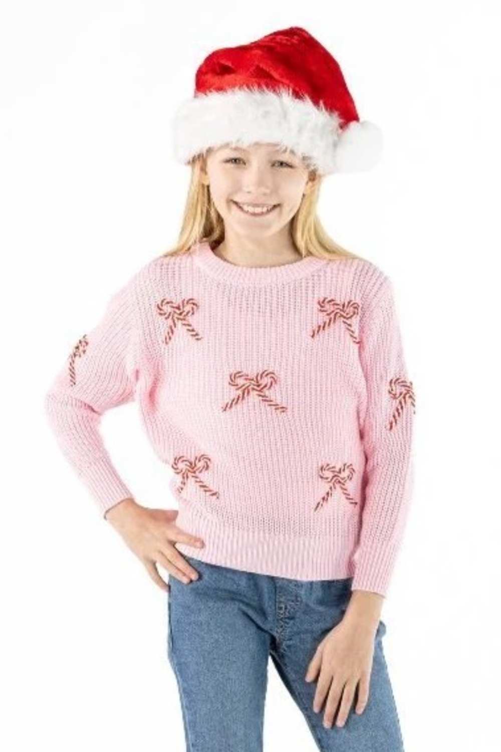 A young girl wearing a pink ribbed knit sweater with red candy cane motifs, blue jeans, and a red Santa hat, posing against a solid white background.