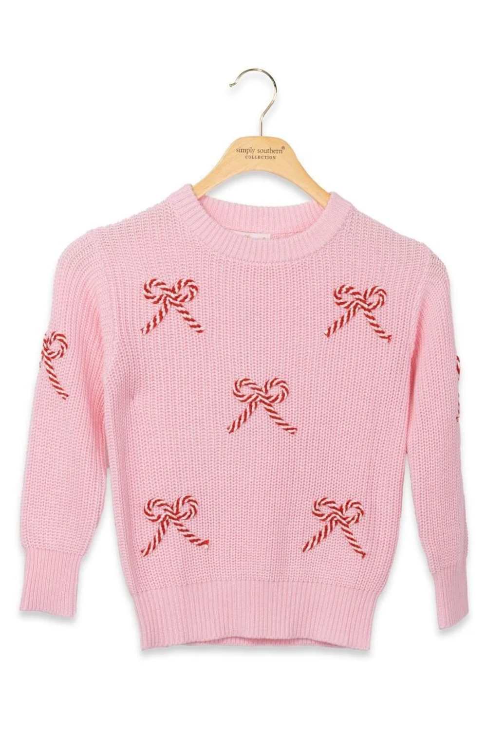 A pink knit sweater with red-and-white candy cane and bow motifs, hanging on a wooden hanger against a plain white background.