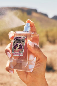 A hand holding a faceted clear perfume bottle with a silver spray nozzle, spraying a fine mist. The bottle features a vintage-style label reading 'Rodeo Soul Waifs'.