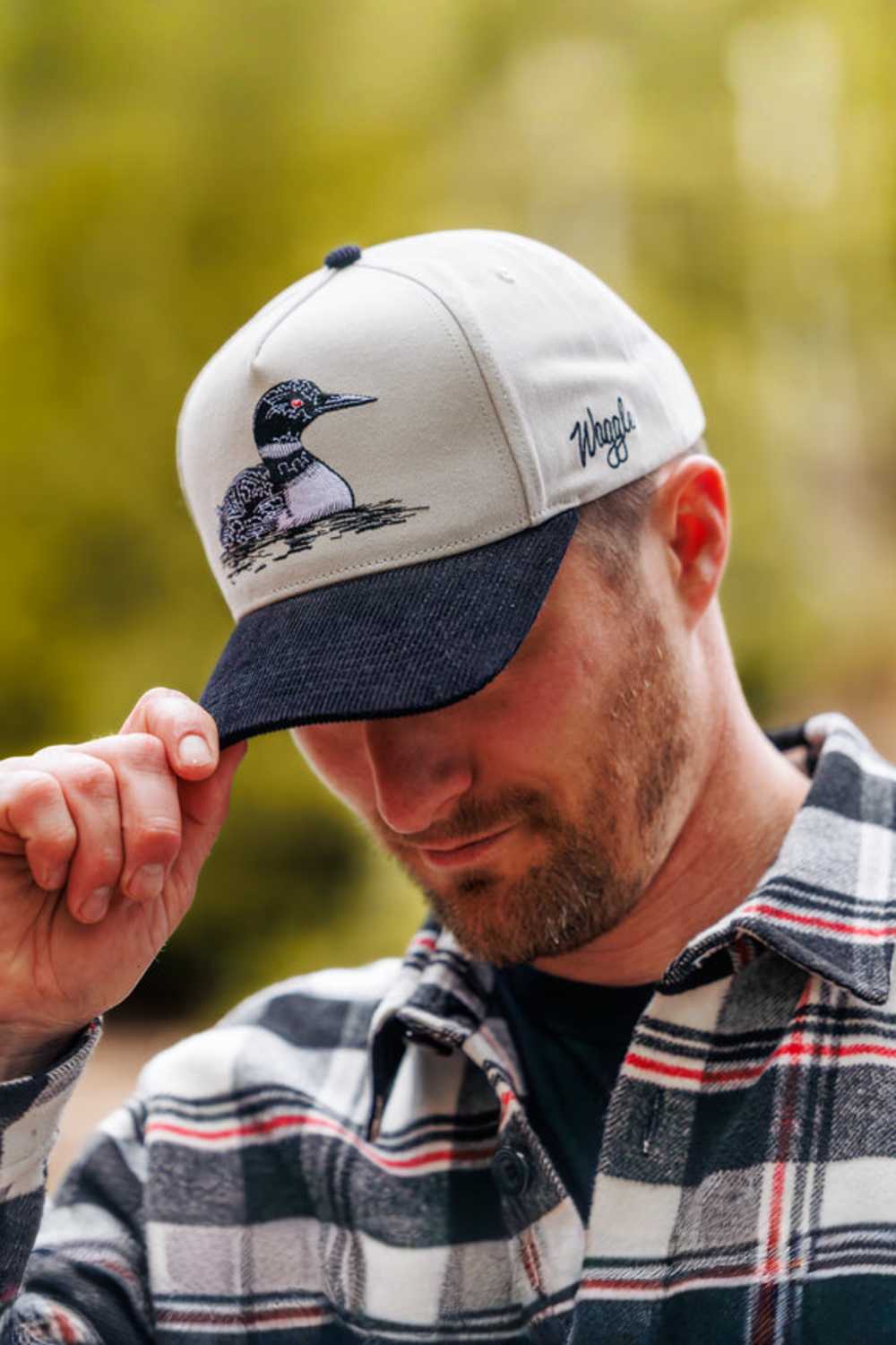 A man wearing a light gray and navy blue baseball cap with an embroidered loon and the brand name on the side, set against a soft-focus green outdoor background.