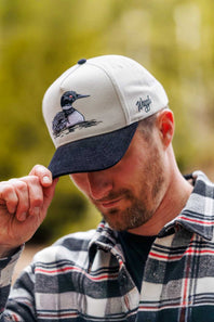 A man wearing a light gray and navy blue baseball cap with an embroidered loon and the brand name on the side, set against a soft-focus green outdoor background.