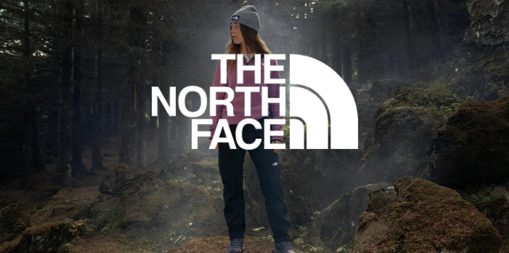 A person wearing a grey beanie, a pink long-sleeve shirt, and dark trousers stands outdoor in a shadowy, misty forest surrounded by mossy rocks and tall trees. The large white THE NORTH FACE logo with its half-dome icon overlaying the center of the image
