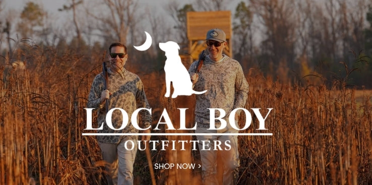Local Boy Outfitters promotional banner featuring two men in camouflage outdoor clothing standing in a field of tall grasses, with the brand’s dog-and-moon logo and a Shop Now call to action