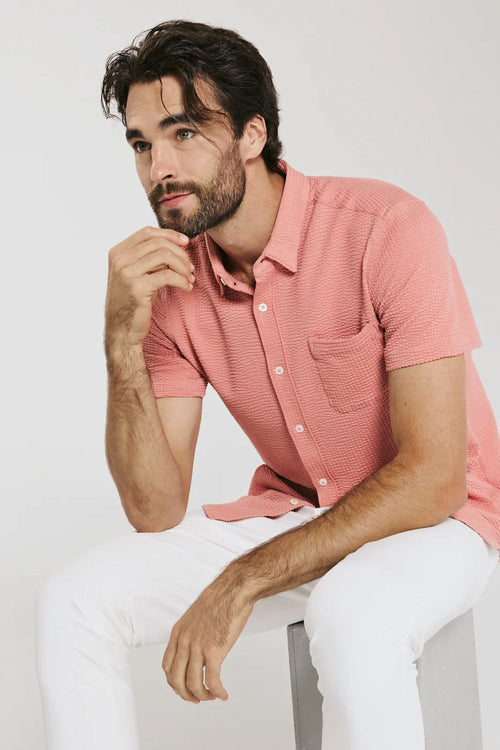 Man wearing a pink shirt and white pants sitting on a white background