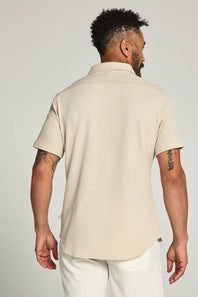 Man wearing a beige polo shirt and white pants against a plain background