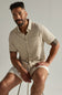 Man wearing a beige shirt and shorts sitting on a stool against a plain background