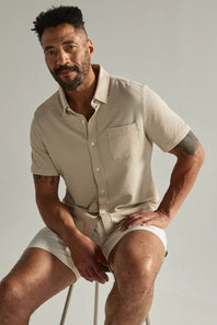 Man wearing a beige shirt and shorts sitting on a stool against a plain background