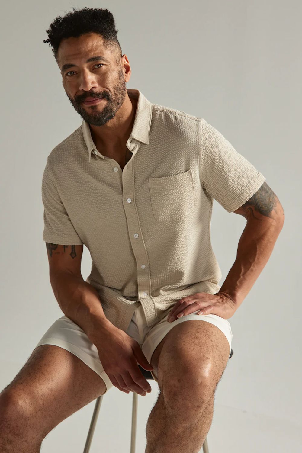 Man wearing a beige shirt and shorts sitting on a stool against a plain background