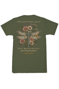 An olive green short-sleeve t-shirt with a detailed graphic on the back. The graphic features a dragonfly made of stained glass flowers and leaves, surrounded by pink and orange wildflowers, with the text 'God turns broken pieces into masterpieces give him your pieces' and 'empty souls.