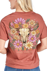 A woman shown from the back wearing a rust-colored short-sleeve t-shirt with a large graphic print of a steer skull surrounded by pink, yellow, and white flowers. The text 'simply southern' is printed below the graphic.