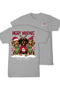 A light gray short-sleeve t-shirt with a festive Christmas graphic of three dachshunds wearing Santa hats and sweaters, with the text 'MERRY WOOFMAS' and 'simply southern' printed below. A second view of the plain gray shirt is shown behind it on a white background.