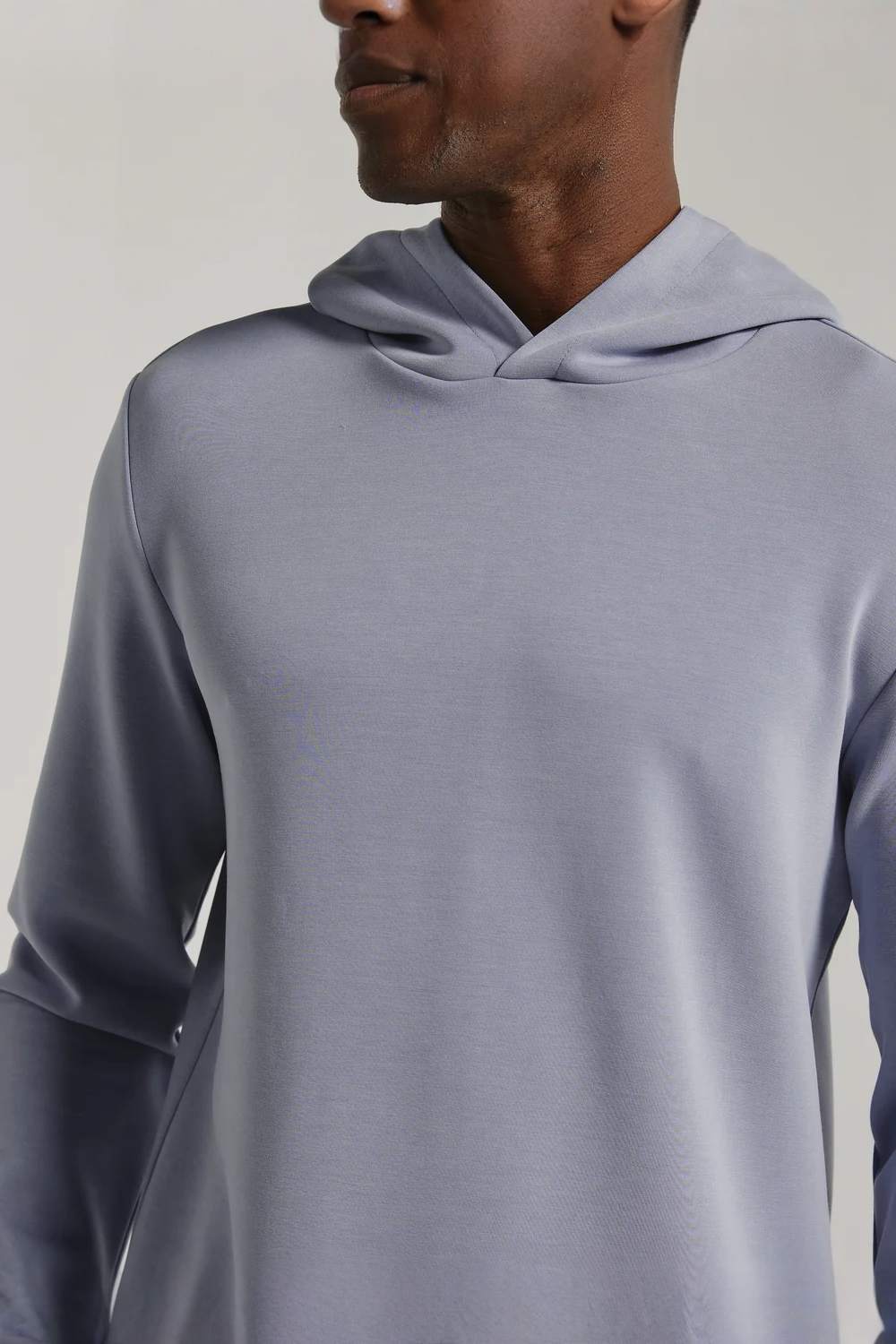 A close-up of a man wearing a light grayish-blue hooded sweatshirt, showing the smooth fabric texture and draped hood against a neutral light gray background.
