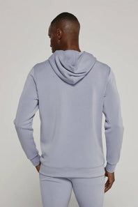 A man viewed from the back wearing a light periwinkle blue hoodie and matching sweatpants against a plain light gray studio background.