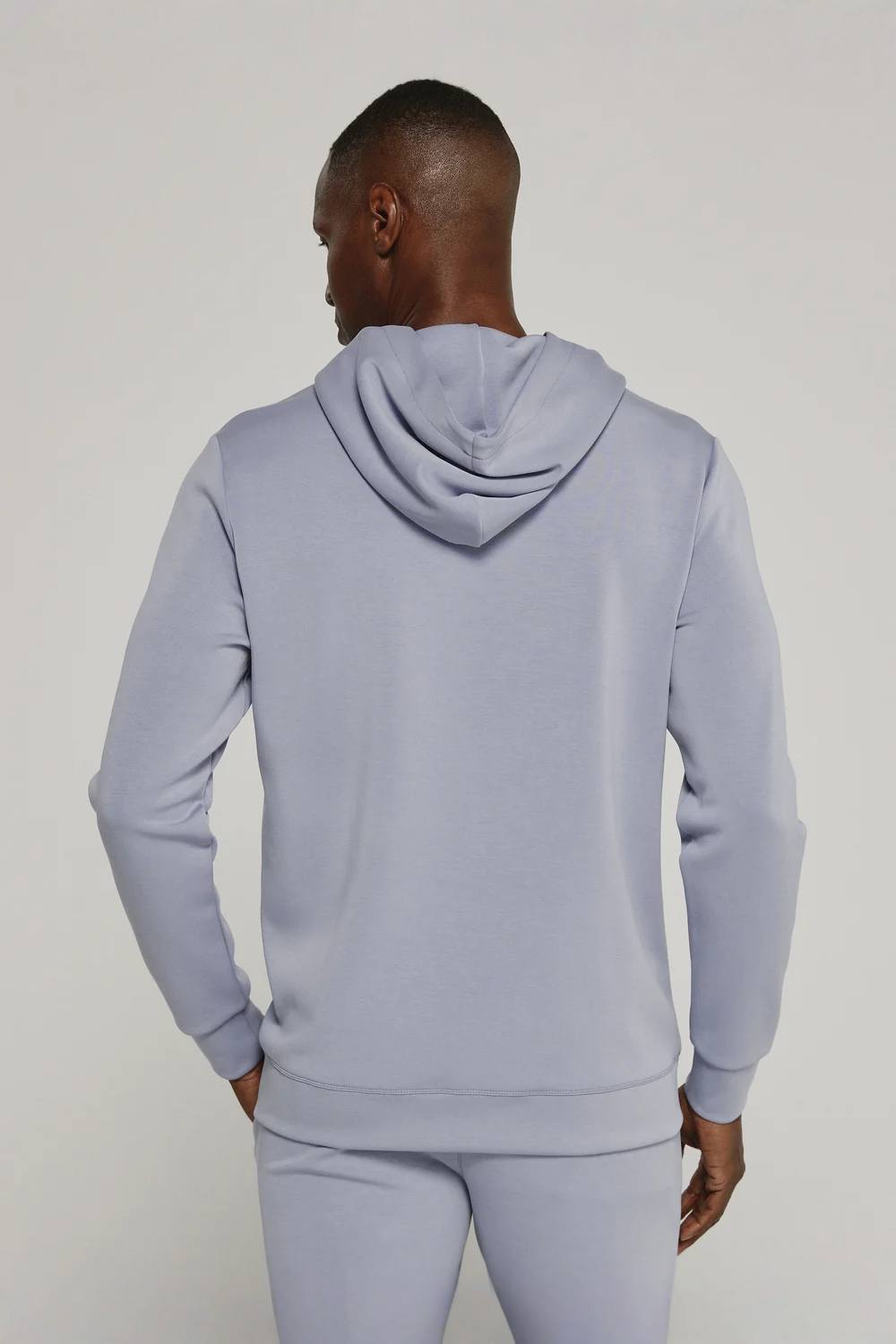 A man viewed from the back wearing a light periwinkle blue hoodie and matching sweatpants against a plain light gray studio background.