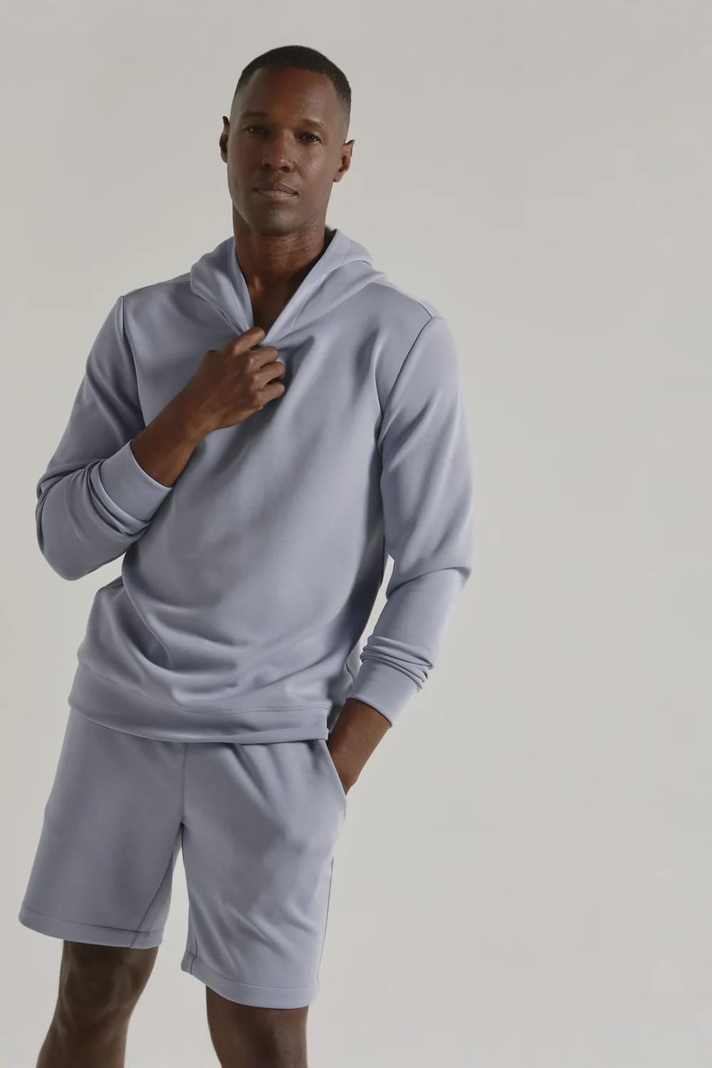 A man modeling a light blue-gray matching loungewear set consisting of a long-sleeve hoodie and matching shorts, photographed against a neutral light gray studio background.
