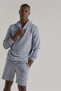A man modeling a light blue-gray matching loungewear set consisting of a long-sleeve hoodie and matching shorts, photographed against a neutral light gray studio background.
