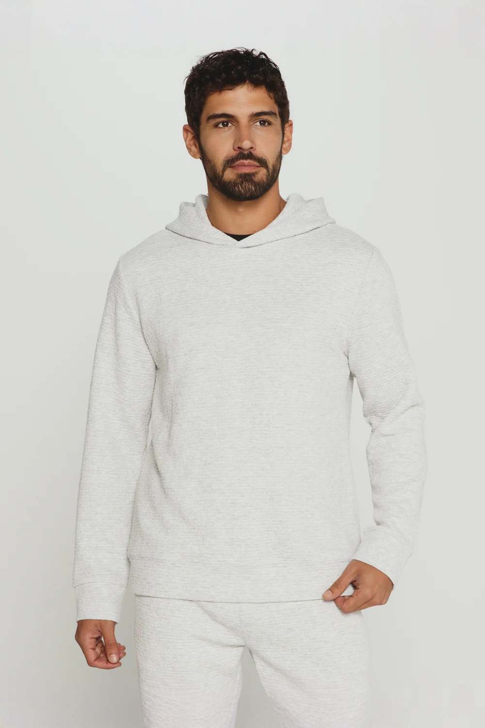 A man wearing a light heather gray hooded sweatshirt and matching sweatpants, standing against a plain light gray studio background.