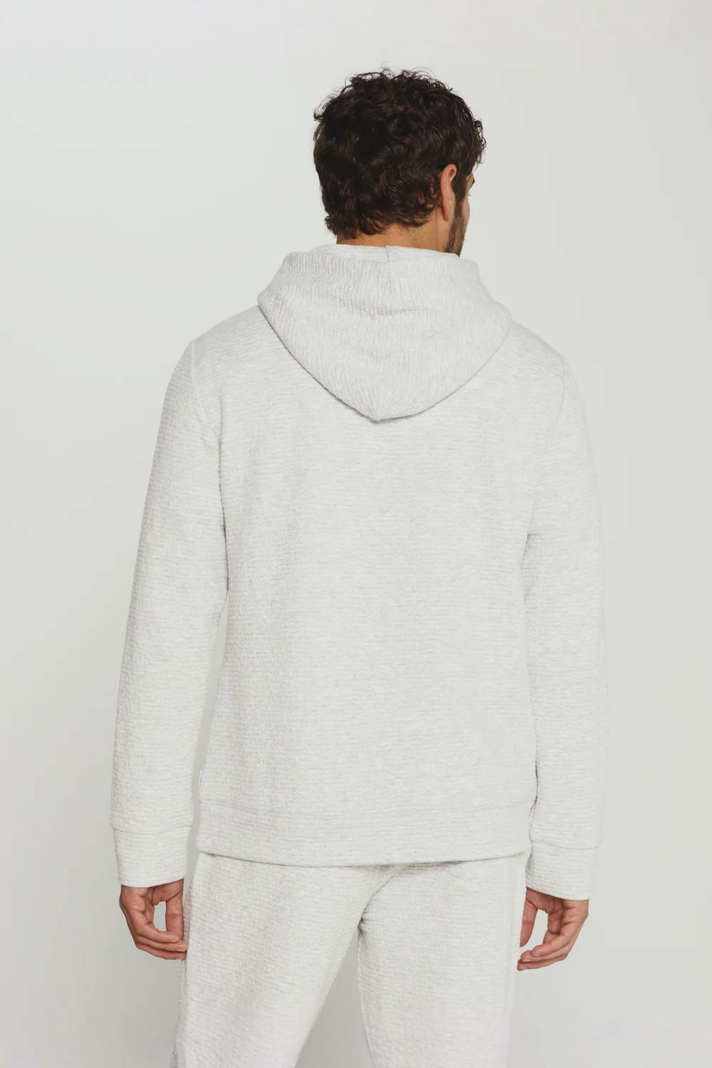 A man viewed from behind wearing a light heather gray hooded sweatshirt and matching sweatpants against a plain light gray studio background.