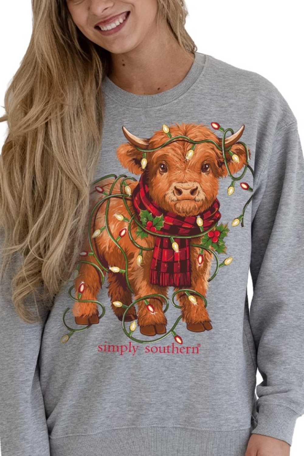 A woman wearing a heather gray crewneck sweatshirt with a graphic of a brown Scottish Highland cow wearing a red plaid scarf and holiday lights, with the text 'simply southern' below.