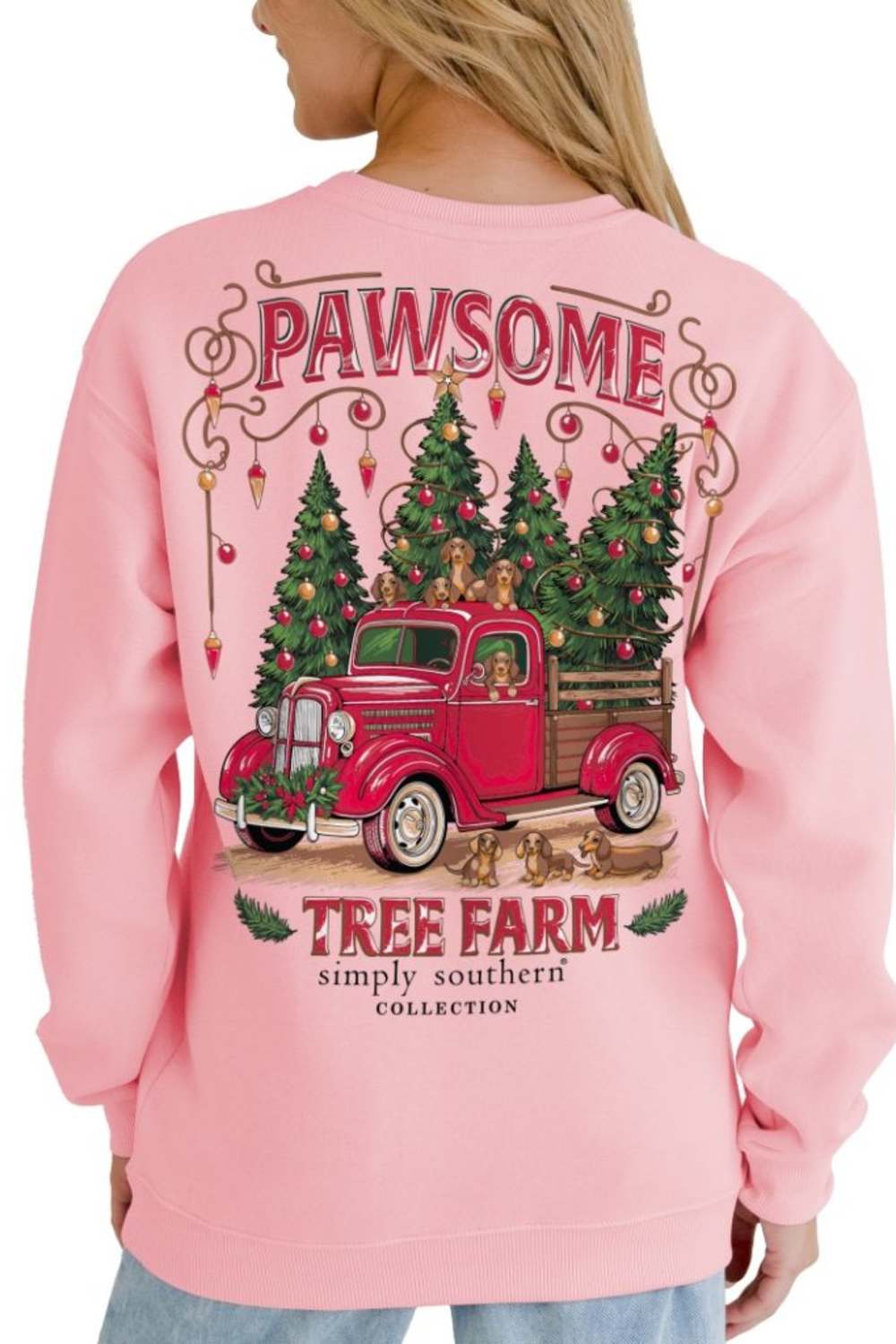 A woman wearing a light pink crewneck sweatshirt, viewed from the back. The sweatshirt features a large graphic print of a red vintage pickup truck carrying Christmas trees, with the words 'PAWSOME' and 'TREE FARM simply southern COLLECTION' printed around it.