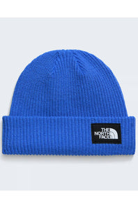 A close-up of a blue ribbed knit beanie hat with a black rectangular patch on the cuff featuring the white 'The North Face' logo, set against a light gray background.
