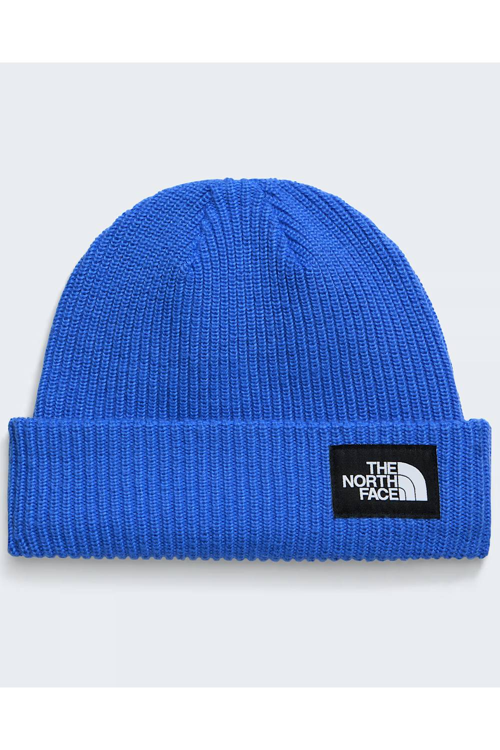 A close-up of a blue ribbed knit beanie hat with a black rectangular patch on the cuff featuring the white 'The North Face' logo, set against a light gray background.