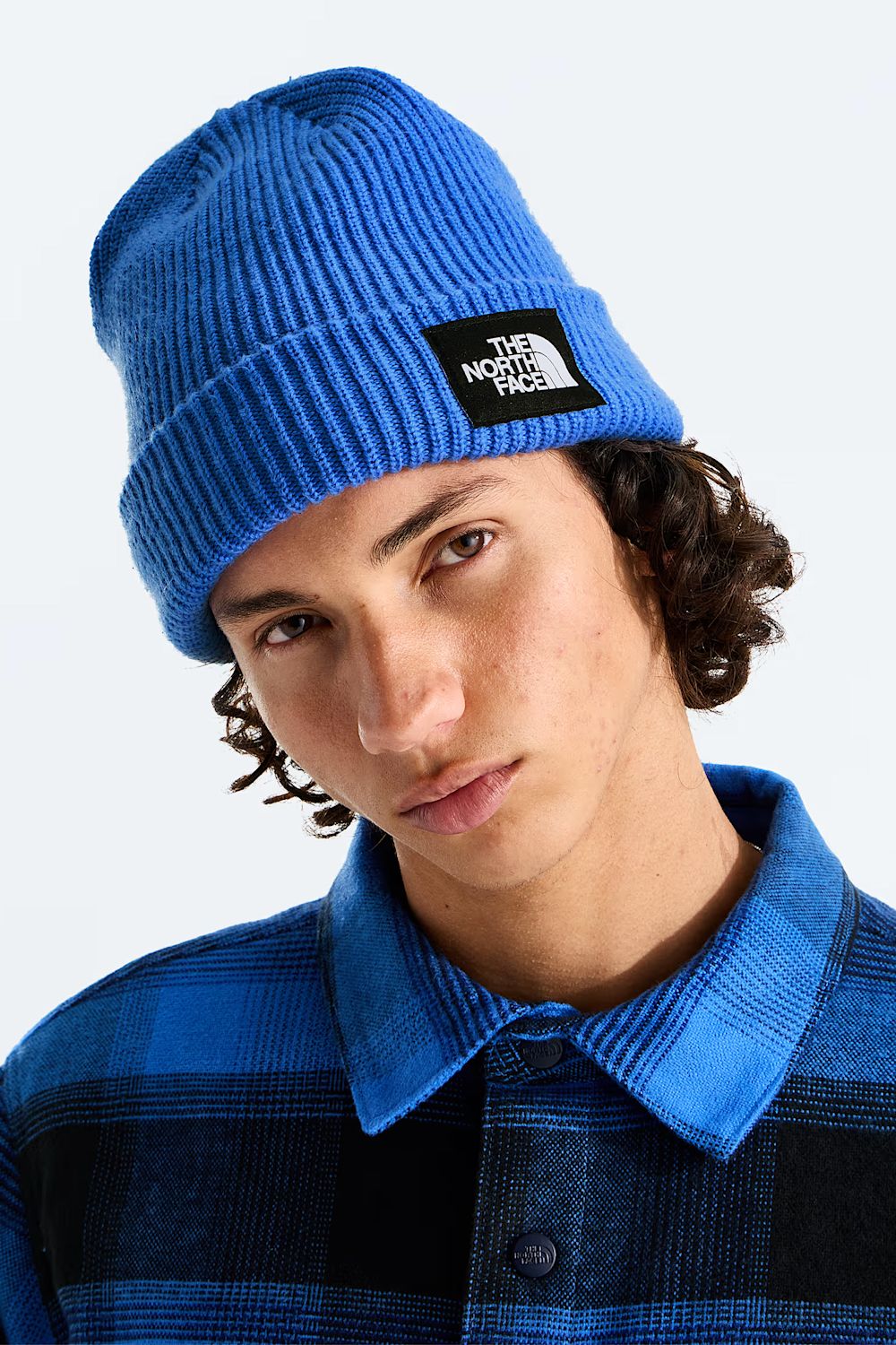 A close-up of a person wearing a bright blue ribbed knit beanie with a black square patch that reads The North Face, paired with a blue and black plaid collared shirt against a light gray background.