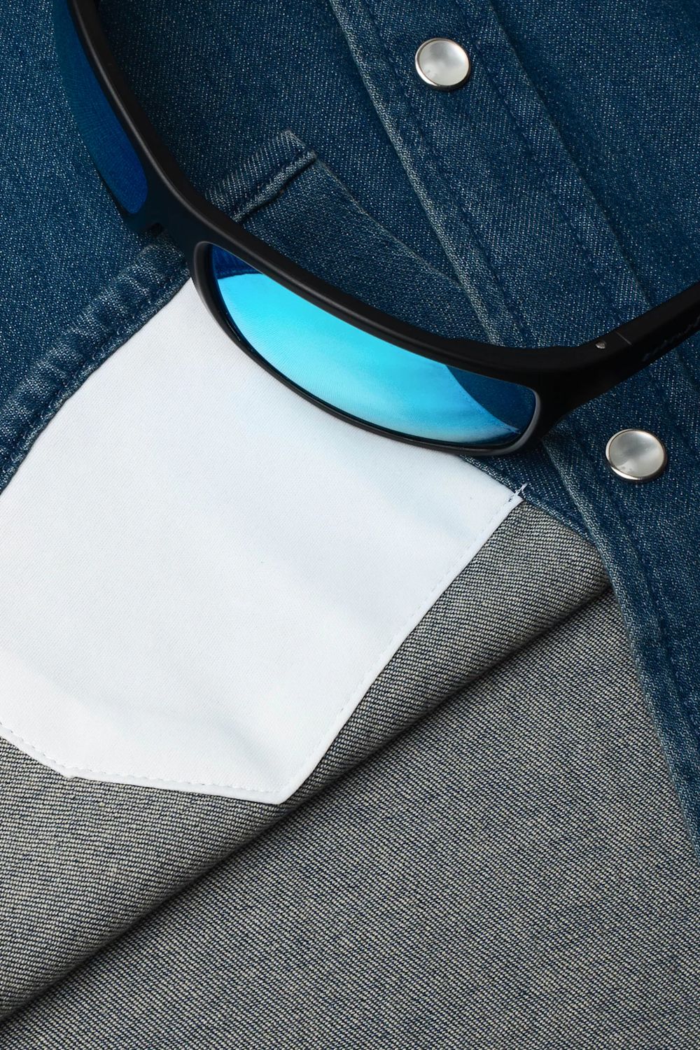 Close-up of a pair of black sunglasses with blue mirrored lenses resting on a folded denim shirt over a gray fabric background.