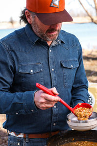 A man wearing a dark blue denim long-sleeve shirt and an orange-brown baseball cap is shown outdoors, using a red ladle to serve food from a large pot into a bowl.