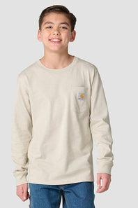 A smiling young boy wearing a light beige long-sleeve pocket t-shirt with a Carhartt logo on the chest, paired with blue jeans, against a solid light gray background.