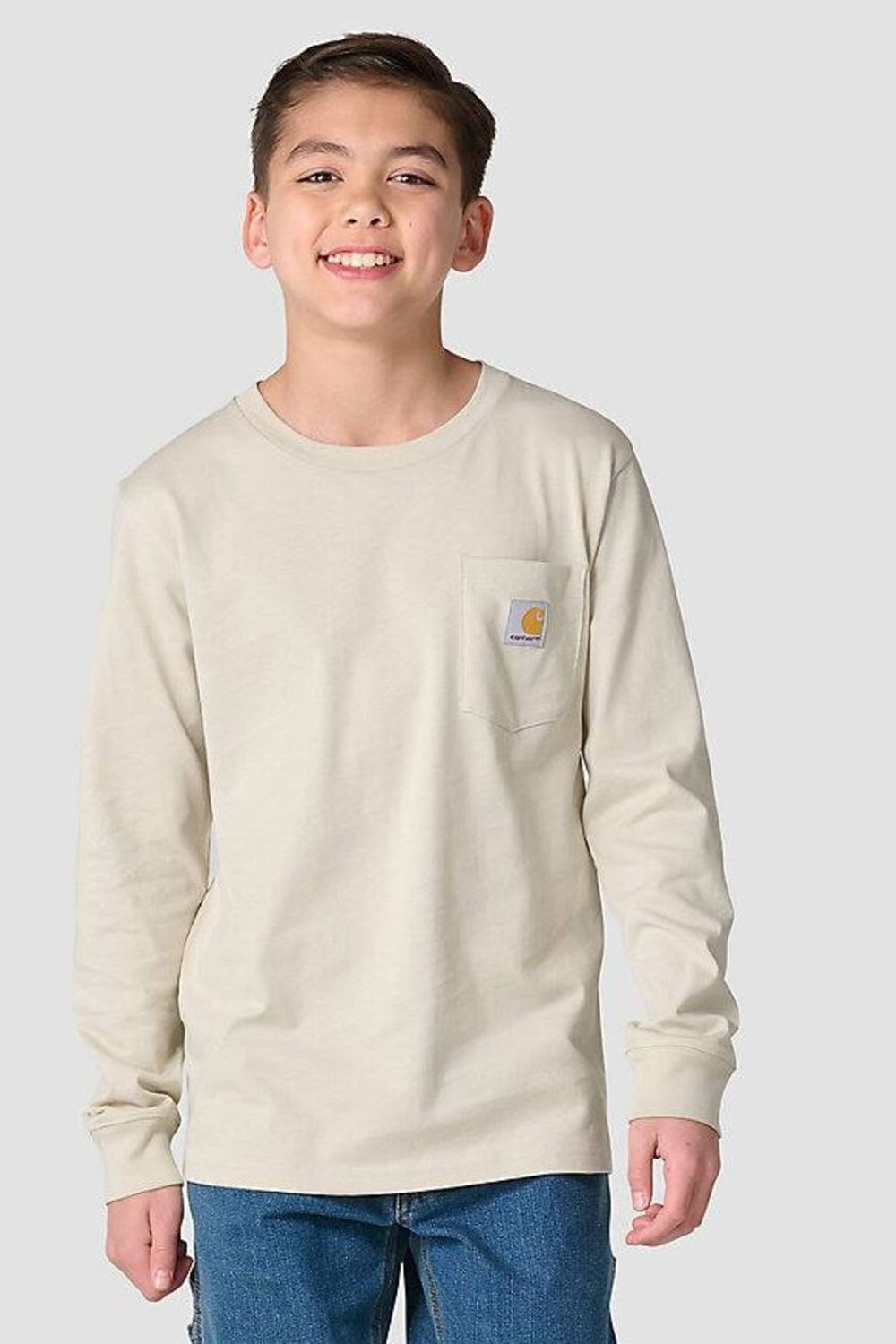 A smiling young boy wearing a light beige long-sleeve pocket t-shirt with a Carhartt logo on the chest, paired with blue jeans, against a solid light gray background.