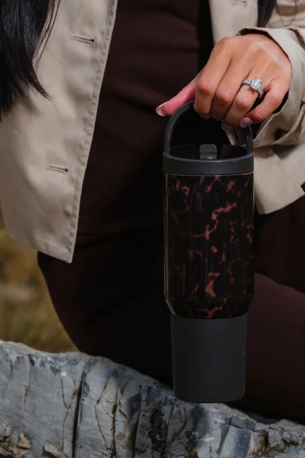 A person outdoors holds a handled travel tumbler with a dark marbled pattern and a matte black base and lid. The person is wearing a beige jacket and dark brown outfit, with a gray stone surface in the foreground.
