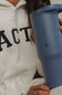 A person in a light gray hoodie holds a matte slate blue HydroJug travel tumbler with a matching lid and carry loop.