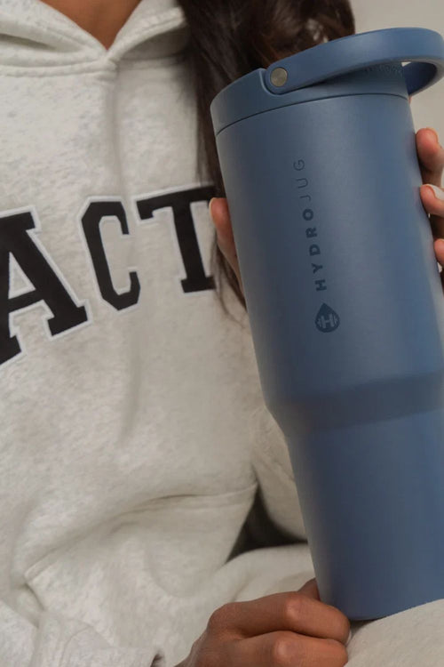 A person in a light gray hoodie holds a matte slate blue HydroJug travel tumbler with a matching lid and carry loop.
