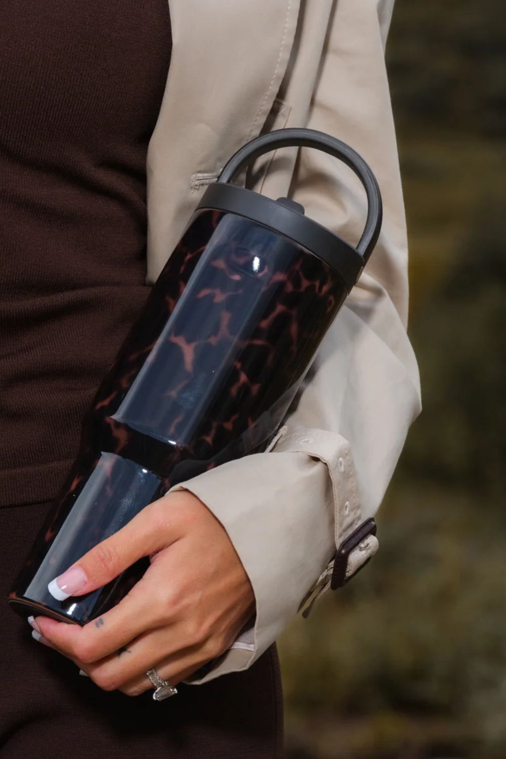 A person holding a cylindrical insulated tumbler with a glossy tortoiseshell pattern in dark brown and black, featuring a black lid and handle, against a blurred outdoor background.