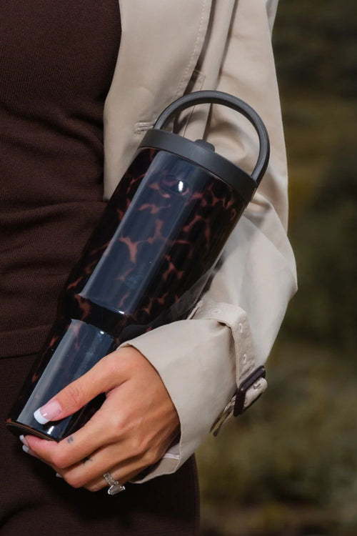 A person holding a cylindrical insulated tumbler with a glossy tortoiseshell pattern in dark brown and black, featuring a black lid and handle, against a blurred outdoor background.