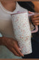 A person holding a tall insulated travel mug with a floral pattern. The mug has a white base with small pink, orange, and green flowers, a light pink lid, and a light pink handle.