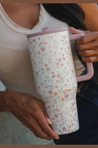 A person holding a tall insulated travel mug with a floral pattern. The mug has a white base with small pink, orange, and green flowers, a light pink lid, and a light pink handle.