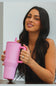 A woman holding a large pink HydroJug tumbler with a handle and straw, shown in a lifestyle setting against a light gray background.