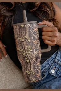 A close-up of a person holding a tall handled tumbler with a realistic woodland camouflage pattern. The tumbler has a gray handle and lid, and the person is wearing a black top and blue denim jeans.