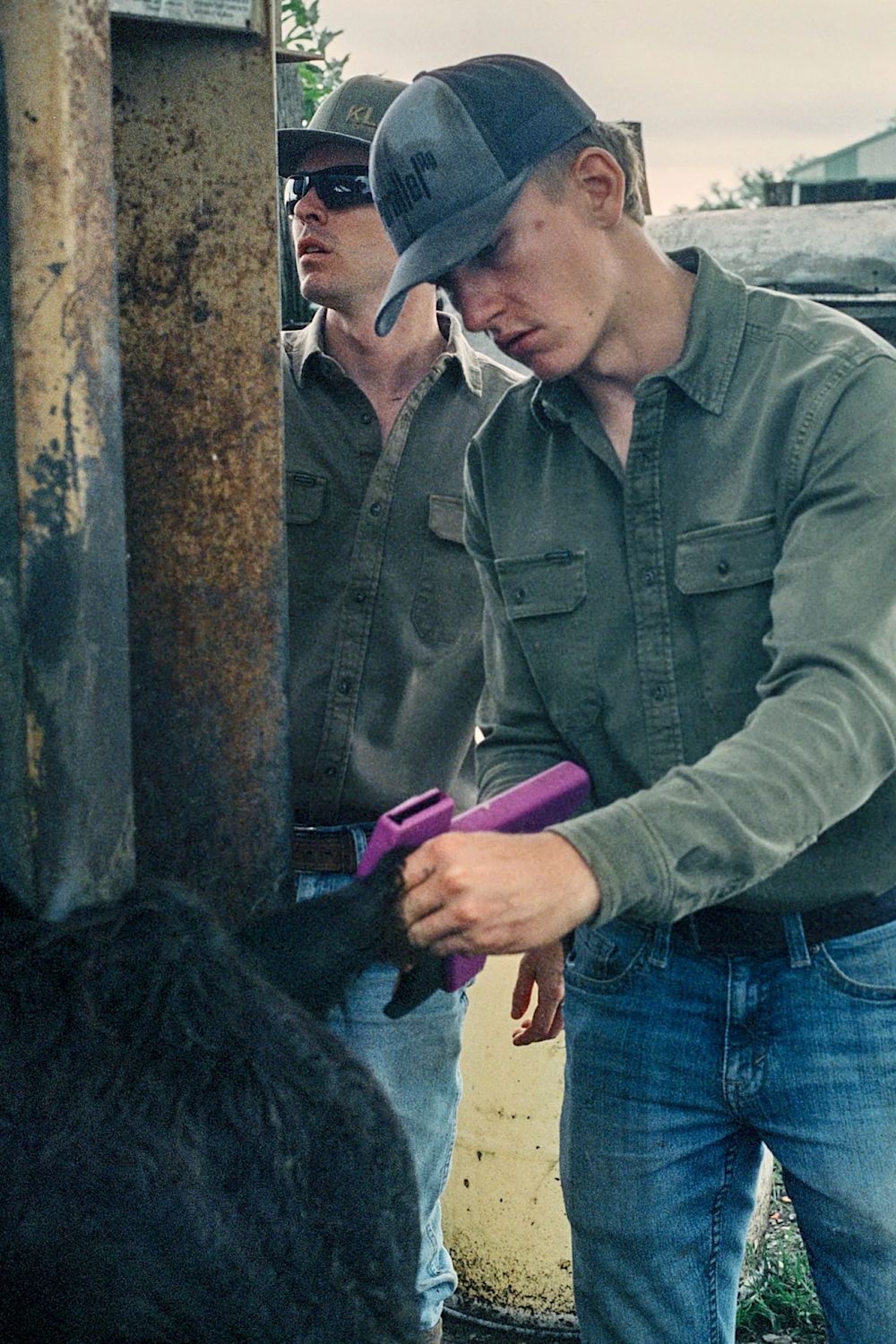A man in a green button-up shirt and baseball cap holds a purple tranquilizer gun while another man in sunglasses stands behind him near a rusty metal post.