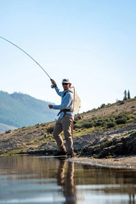 A man fly fishing in a river, casting a rod with a bent line, wearing a light blue long-sleeve shirt, khaki pants, and a white cap, with a backpack visible behind him against a mountain and sky background.